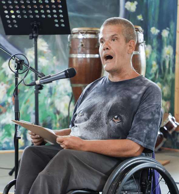 A man in a wheelchair speaking into a microphone. His shirt has the eyes of an animal on it.