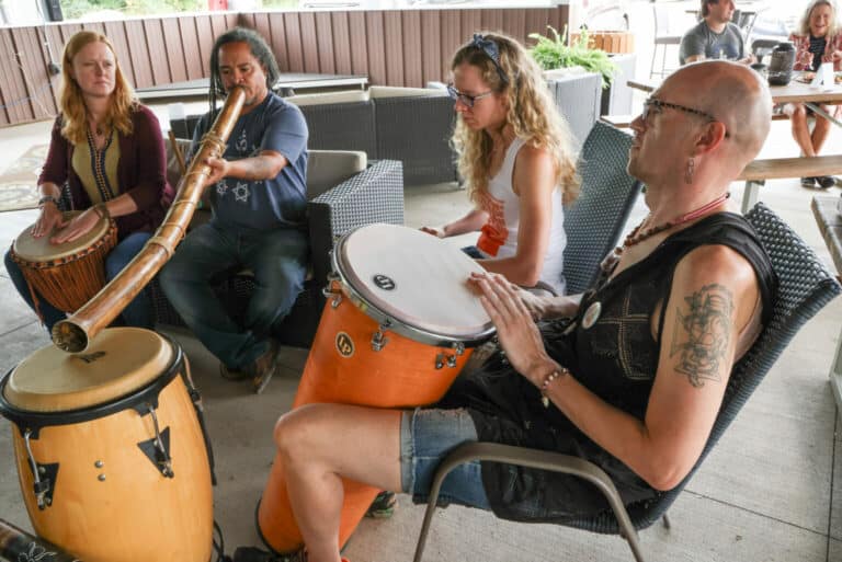 A group of four adults in a drum circle. They sit in chairs. One plays a didgeridoo while the rest play large drums with their hands.